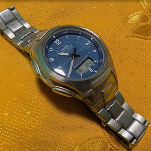 May include: A silver-toned Casio watch with a blue face and digital display. The watch features a stainless steel band and a round face with white numerals and hands. The watch is displayed on a gold-colored surface.