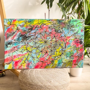 May include: Abstract art painting on canvas. The artwork features a colorful background with red, yellow, blue, and green hues. A black geometric design is overlaid on the colorful background. The painting is displayed on a wooden easel.