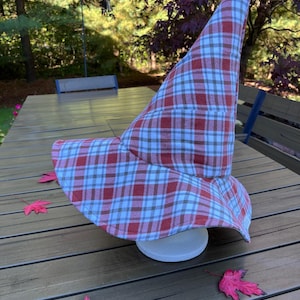 May include: A pointed hat featuring a plaid pattern in shades of red, white, and dark gray. The hat has a wide, slightly curved brim and a tall, conical crown, constructed from a soft fabric material that maintains a structured shape.