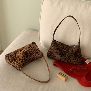 Może przedstawiać: Dwie torebki w panterkę z brązowymi i czarnymi plamami. Jedna torba ma długi uchwyt, druga krótszy. Leżą na jasnej powierzchni z czerwonym swetrem, okularami i małym pudełkiem.