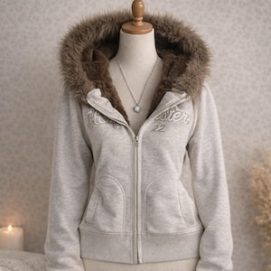 Può includere: Una felpa con cappuccio grigio chiaro con zip e cappuccio foderato in finta pelliccia. La felpa ha la scritta "Hollister" ricamata in bianco, con il numero "22" sotto. È visibile una collana d'argento con un ciondolo.
