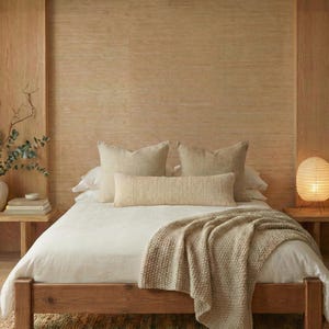 May include: A bedroom with a wooden bed frame, white duvet, and beige pillows. A beige knitted throw blanket is on the bed. The room has light-coloured wooden walls and a woven rug.