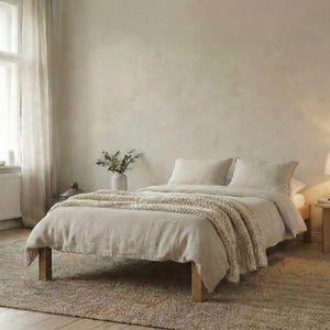 May include: A bedroom in neutral tones featuring a bed, rug, and window. The bed has a beige duvet and pillows, with a knitted throw. A small wooden side table holds a lamp. The room has a natural, minimalist aesthetic.