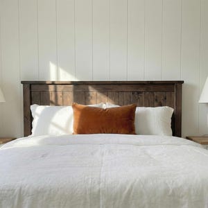 May include: A bed featuring a dark brown wooden headboard, white bedding, and an orange cushion. Two white lamps flank the bed. The wall behind the bed is white with vertical wooden panels.