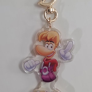 Può includere: Un portachiavi in acrilico trasparente con un personaggio dei cartoni animati con capelli biondi, un naso grande e un abito rosso e viola. Il portachiavi ha un fermaglio e un anello in metallo dorato.
