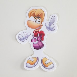 Può includere: Un adesivo fustellato con il personaggio Rayman. Il personaggio ha i capelli biondi, un naso grande e indossa una felpa con cappuccio rossa e pantaloni viola. Le sue mani sono bianche e le sue scarpe sono gialle e bianche. L'adesivo è su sfondo bianco.