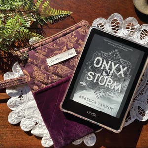 Può includere: Un e-reader Kindle che mostra il libro "Onyx Storm" di Rebecca Yarros, accanto a una custodia per libri trapuntata e fantasia, viola. La custodia ha un cinturino da polso e un'etichetta che dice "The Whisper Page". Gli oggetti poggiano su una tovaglietta di pizzo bianco.