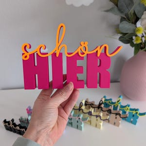 Könnte beinhalten: Ein leuchtend rosa Holzschild mit den deutschen Worten "schön hier" in orangefarbener Schrift. Das Schild wird hochgehalten, mit mehreren kleineren Schildern in verschiedenen Pastellfarben darunter. Der Hintergrund umfasst eine Blumenvase.