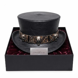 May include: A black top hat with a textured finish, featuring a band decorated with a brown snakeskin pattern, silver-toned metal skull accents, and studs. A small rectangular card in front displays the text FOR A CONNOISSEUR in black lettering.