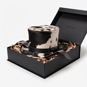 May include: A top hat with a black and white cowhide pattern, featuring a black leather band around the crown. The hat rests inside a black rectangular box labeled STETSON, nestled among brown paper filler strips with a grey ribbon accent.