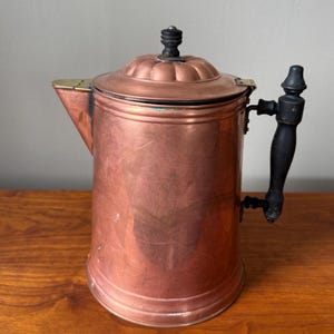 May include: A copper coffee pot with a black handle and lid finial. The pot has a spout and a hinged lid. The copper has a warm, reddish-brown color, and the handle and lid finial are black. The pot is likely vintage.