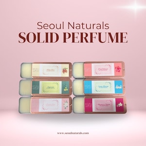 May include: Solid perfume tins from Seoul Naturals, in a variety of scents. Each rectangular tin has a bronze-colored lid and contains a solid perfume balm. The tins are labeled with the scent name and a small graphic. The background is a soft pink.