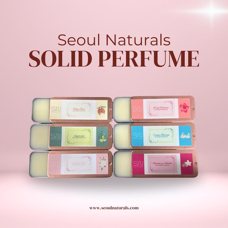 May include: Solid perfume tins from Seoul Naturals, in a variety of scents. Each rectangular tin has a bronze-colored lid and contains a solid perfume balm. The tins are labeled with the scent name and a small graphic. The background is a soft pink.