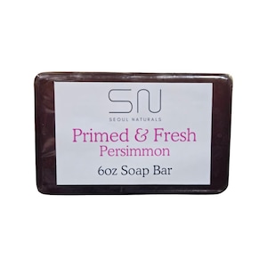 Op de afbeelding: Een donkerbruin rechthoekig stuk zeep met een wit etiket. Het etiket toont de tekst "Primed & Fresh Persimmon" en "6oz Soap Bar". De merknaam "Seoul Naturals" is ook zichtbaar.