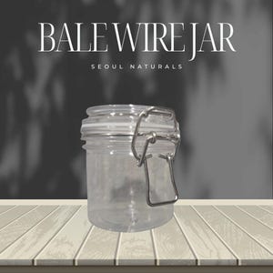 May include: Clear glass jar with a metal bale wire closure. The jar has a round shape and a clear lid. The text "BALE WIRE JAR" and "SEOUL NATURALS" are displayed in white letters above the jar.