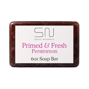 May include: A rectangular 170g soap bar with a dark reddish-brown translucent appearance. A white label in the centre features the black text SN SEOUL NATURALS, the pink text Primed & Fresh Persimmon, and the black text 6oz Soap Bar.