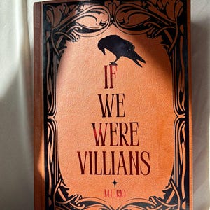 May include: A hardcover book with a burnt orange cover featuring the title "IF WE WERE VILLIANS" in red lettering. A black raven silhouette sits above the title. The cover is framed by an ornate black design.