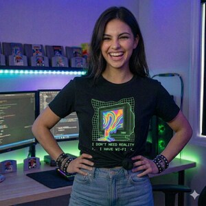 Op de afbeelding: Zwart t-shirt met een retro computer graphic en de tekst "I DON'T NEED REALITY > I HAVE WI-FI <". Het computerscherm toont een kleurrijk, smeltend ontwerp. Het t-shirt wordt gedragen met een blauwe jeans.