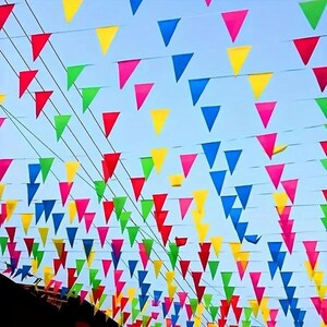 May include: A series of triangular pennant flags in vibrant shades of red, yellow, blue, green, and pink, suspended on multiple parallel strings against a clear light blue sky, creating a festive overhead canopy of colorful geometric shapes.