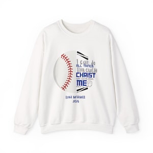 Peut inclure: Un sweat-shirt blanc à manches longues avec un motif de balle de baseball aux coutures rouges. Le texte indique I CAN DO ALL THINGS THROUGH CHRIST WHO STRENGTHENS ME, PHILIPPIANS 4:13, LAKE BASEBALL et 2026, avec deux battes noires et un gant.
