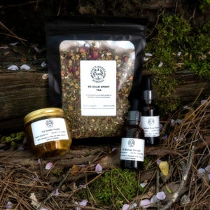 Può includere: Un assortimento di prodotti naturali per il benessere, tra cui un barattolo di miele, una busta di tè e due flaconi di tinture. I prodotti sono disposti su un letto di foglie e ramoscelli, con un tronco di legno sullo sfondo. Le etichette sono visibili.