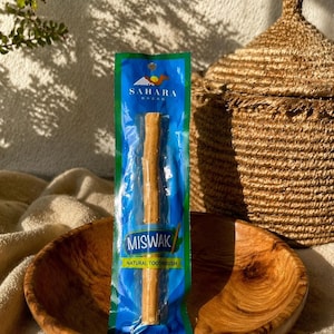 Puede incluir: Un cepillo de dientes natural Miswak empaquetado, presentado en un cuenco de madera. El embalaje es azul y verde con el texto "Sahara Bazar" y "Miswak". Una cesta tejida se encuentra en el fondo.