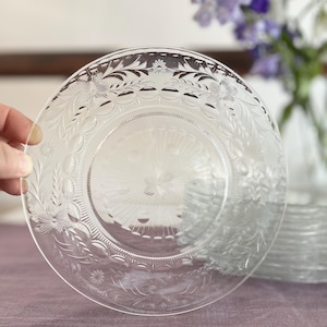 Puede incluir: Plato de vidrio transparente con diseño floral y de hojas grabado. El plato está sostenido por una mano y tiene un patrón detallado alrededor del borde y el centro. Una pila de platos similares es visible en el fondo.
