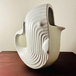 Può includere: Una brocca in ceramica bianca con un design scultoreo unico. La brocca presenta un manico e un beccuccio. La superficie è strutturata con motivi a onde. La brocca è di un colore chiaro e neutro.