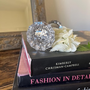 May include: A stack of three books with the titles "Fashion in Detail" and "Fashioning Fashion" is topped with a clear, faceted glass doorknob and white flowers. The books are on a wooden surface.