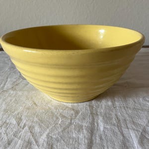 Peut inclure: Un bol en céramique jaune avec un extérieur nervuré. Le bol a une forme arrondie et une finition lisse et brillante. Le bol est posé sur une surface blanche et texturée.