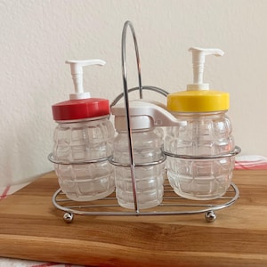 May include: Three clear glass dispensers with a textured, grid-like pattern, each featuring a white pump mechanism. The left dispenser has a red lid, the center has a white lid, and the right has a yellow lid. They are held in a chrome wire caddy.