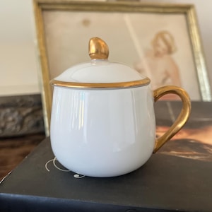 May include: A white ceramic mug with a gold handle and trim, topped with a matching lid. The mug is sitting on a book with the text "KIMBERLY CHRISMAN-CAMPBELL". The mug has a rounded shape and a gold knob on the lid.
