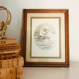 May include: A framed illustration of a frog wearing a coat and hat while fishing in a pond with a rod and line. The artwork is enclosed in a rectangular wooden frame with a light green mat border, featuring a circular central image.