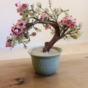 May include: A decorative bonsai tree sculpture featuring a textured brown trunk, translucent pale green leaves, and clusters of small pink and white blossoms. The tree is set in a light green ceramic pot filled with small white pebbles.