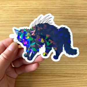 May include: A holographic sticker of a black cat with a startled expression. The cat's fur is a mosaic of dark blue and iridescent colors, with yellow accents on its paws. The sticker is held against a light wood grain surface.
