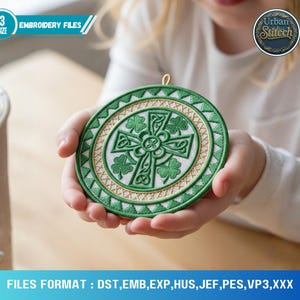 May include: A green and gold embroidered round design featuring a Celtic cross and shamrocks. The design is held in a person's hands. The text "EMBROIDERY FILES" and "Urban Stitch" are visible. The design is approximately 4 inches in diameter.
