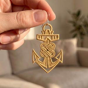 May include: A gold-colored embroidered anchor earring featuring a central knot design and a silver-toned metal hook. The anchor is constructed from dense, textured thread work with a geometric pattern at the top crossbar and pointed flukes.