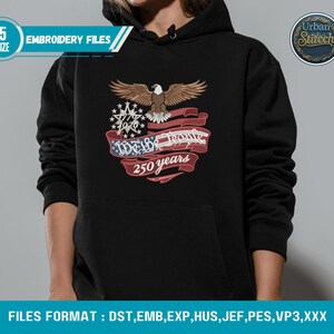 K&ouml;nnte beinhalten: Schwarzer Kapuzenpullover mit einem gestickten Design mit einem Adler, einem Banner mit "We the People 250 years" und einem Schneeflocken-Design. Der Hoodie hat eine Vordertasche und eine Kapuze mit Kordelzug. Der Text "EMBROIDERY FILES" ist ebenfalls sichtbar.