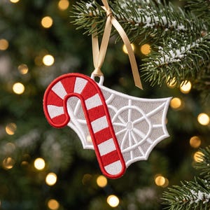 May include: A holiday ornament featuring a red and white striped candy cane layered over a white embroidered spider web design. The ornament is constructed from stitched fabric and hangs from a thin, light gold ribbon loop for display.