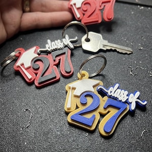 May include: Three keychains featuring a graduation cap and the text "class of 27". One keychain is red with a black "27", one is yellow with a blue "27", and one is red with a white cap. Each includes a metal ring for keys.