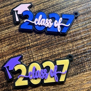 May include: Two 3D-printed graduation ornaments featuring the text "class of 2027". The top ornament is blue with a light pink graduation cap and script text. The bottom ornament is yellow with a purple graduation cap and script text.
