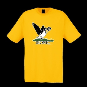 May include: Mustard yellow t-shirt featuring a graphic of a bird with black wings and a white body, holding a pretzel. The word "IRONIC" is printed below the bird in white. The shirt is a short-sleeved crew neck.