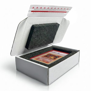 May include: White cardboard box with a black foam insert, designed to hold a trading card. The box is open, revealing the card inside a protective case. The top flap has red markings.