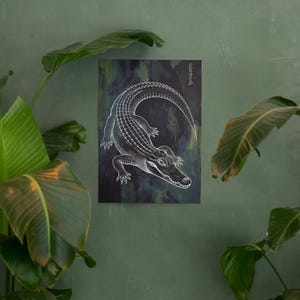 May include: White line art illustration of an alligator on a dark, textured background. The artwork is mounted on a rectangular canvas. Japanese characters are visible in the upper right corner. The artwork is displayed against a green wall with leafy plants.