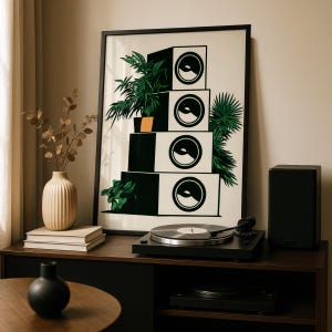 May include: A framed wall art print featuring a vertical stack of four white square speakers with black circular cones, accented by green leafy plants on the sides, displayed in a black frame on a dark wooden surface.