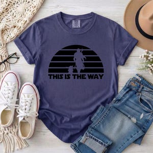 May include: A navy blue t-shirt featuring a black silhouette design of a figure and a child against a striped sunset. The text "THIS IS THE WAY" is printed below the design. The shirt is displayed with white sneakers and blue jeans.