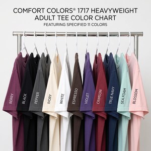 May include: A color chart for Comfort Colors 1717 heavyweight adult tees featuring 11 shirts hanging on white hangers. The colors are labeled as Berry, Black, Pepper, Ivory, White, Espresso, Violet, Crimson, True Navy, Sea Foam, and Blossom.