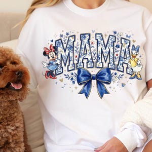 Pode incluir: Camiseta branca com a palavra "MAMA" em letras azuis, decorada com padr&otilde;es florais e um la&ccedil;o azul. Minnie Mouse e Margarida est&atilde;o na camiseta. Um c&atilde;o marrom est&aacute; em primeiro plano.