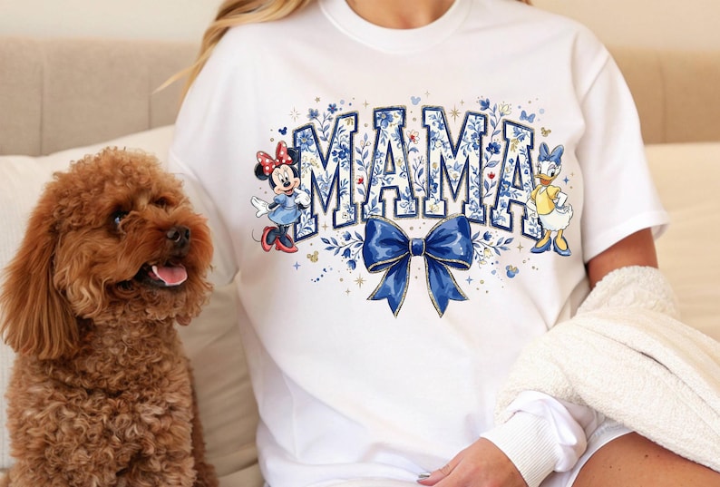 Pode incluir: Camiseta branca com a palavra "MAMA" em letras azuis, decorada com padr&otilde;es florais e um la&ccedil;o azul. Minnie Mouse e Margarida est&atilde;o na camiseta. Um c&atilde;o marrom est&aacute; em primeiro plano.