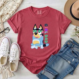 May include: A brick red crew neck t-shirt featuring a graphic of a blue and yellow cartoon dog sitting with crossed legs. The dog has a pink flower crown and the text "GIRL DAD" is printed vertically in blue, pink, and green letters next to it.